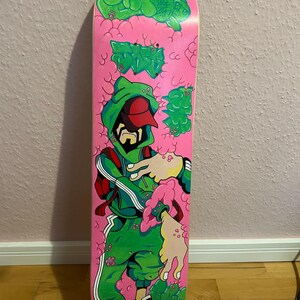 May include: A pink skateboard deck with a green cartoon character wearing a red hat and a green hooded sweatshirt. The character is surrounded by green and pink bubble-like shapes.