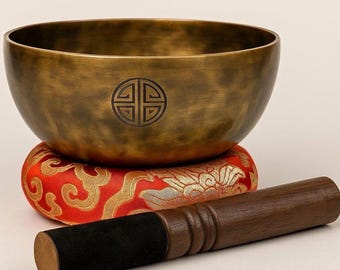 May include: A bronze singing bowl with a carved design, resting on a red and gold cushion. A wooden mallet with a black grip lies beside the bowl. The bowl is a meditation tool.