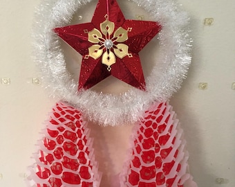 Peut inclure: Décoration de Noël festive avec une étoile rouge à détails dorés, au centre d'une couronne de guirlande argentée. En dessous, deux ornements en papier rouge et blanc suspendus.