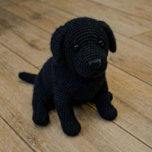 Könnte beinhalten: Ein handgehäkelter schwarzer Labrador-Welpe sitzt auf einem Holzboden. Der Welpe hat ein detailliertes, strukturiertes Aussehen mit sichtbaren Stichen, einem kleinen Schwanz und dunklen Augen. Das Objekt ist handgefertigt.