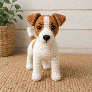 Pode incluir: Uma figura de cão em croché com corpo branco e manchas castanhas nas orelhas, cabeça e costas. O brinquedo tem um nariz preto, olhos redondos escuros e uma cauda curta erguida. A textura é criada com pontos de fio visíveis.