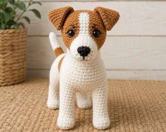 Puede incluir: Una figura de perro tejida a ganchillo con cuerpo blanco y manchas marrones en las orejas, la cabeza y el lomo. El juguete tiene una nariz negra, ojos redondos oscuros y una cola corta erguida. La textura presenta puntos de hilo visibles.