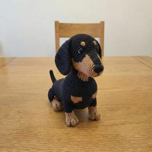 May include: A crocheted dachshund dog in black and tan colours. The dog is positioned on a wooden table, with a wooden chair visible in the background. The dog features floppy ears, a long body, and a small tail.