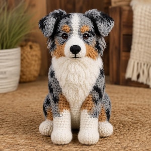 May include: A crocheted stuffed animal in the likeness of an Australian Shepherd dog. The plush features a mottled coat of gray, black, and white yarn with tan patches around the eyes and ears, a white chest, and dark brown plastic eyes.