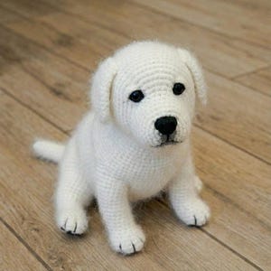 Puede incluir: Un cachorro de perro blanco tejido a ganchillo con ojos y nariz negros, sentado en un suelo de madera. El perro tiene orejas caídas y una cola pequeña. El trabajo de ganchillo es detallado y texturizado.