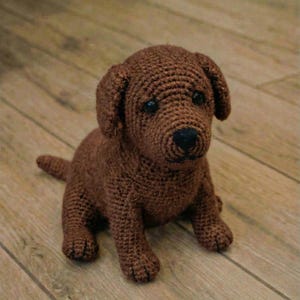 以下が含まれることがあります： 黒い目と鼻を持つ、手作りの茶色の犬の編みぐるみ。犬は木の表面に座っています。子犬は垂れ耳と小さな尻尾を持っています。このぬいぐるみは、子供たちへの素晴らしい贈り物です。