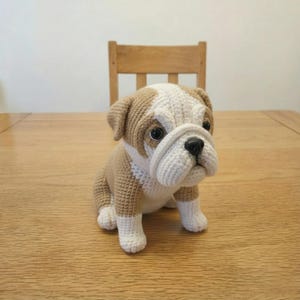 Puede incluir: Un perro bulldog tejido a ganchillo en tonos beige y blanco. El perro tiene una nariz negra y ojos negros. Está colocado sobre una mesa de madera. Una silla de madera es visible en el fondo.