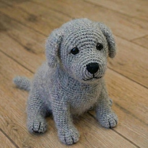Puede incluir: Un cachorro gris tejido a mano, con ojos y nariz negros. El peluche está sentado en un suelo de madera. El perro tiene orejas caídas y una cola pequeña. El perro mide aproximadamente 20 cm de altura.