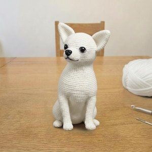 May include: A white crochet Chihuahua dog figurine. The dog is sitting upright with its ears perked up and has black eyes and nose. The figurine is on a wooden table, with a ball of white yarn and crochet hooks in the background.