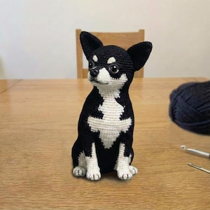 Puede incluir: Figura de perro chihuahua hecha a mano en crochet blanco y negro. El perro está sentado con las orejas erguidas. La figura tiene el pecho y las patas blancas, el resto del cuerpo es negro. El perro está sobre una mesa de madera.