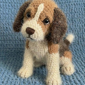 May include: A handmade crochet beagle dog with brown, white, and tan coloring. The plush toy has dark eyes, floppy ears, and a small tail. The dog is sitting on a blue crocheted surface.