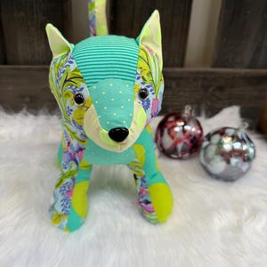 May include: A colorful fabric fox toy with a patchwork design. The fox features a teal striped head, dotted snout, and floral patterned ears and legs. The toy is sitting on a white fluffy surface, with two decorative glass ornaments in the background.