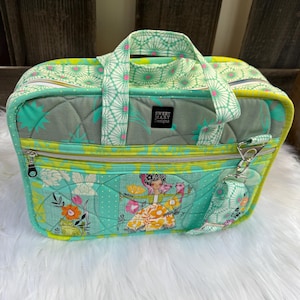 May include: A quilted green and yellow fabric bag with a zipper closure and a handle. The bag has a floral print and a label that says "Sweet Mary Designs".