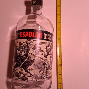 May include: A clear glass Espolon Tequila Blanco bottle with a wooden cork stopper and a black and white label featuring skeleton illustrations. A yellow measuring tape placed next to the bottle indicates a height of approximately 12 inches.