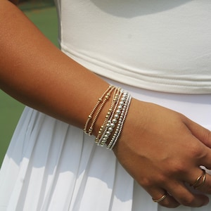 May include: A stack of five beaded bracelets worn on the wrist, featuring three gold-toned bracelets with varying bead sizes and two silver-toned bracelets with uniform beads. Two simple gold-toned rings are worn on the fingers of the same hand.