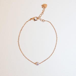 May include: A delicate gold chain bracelet with a small, clear gemstone in the centre. The bracelet has a heart-shaped clasp.