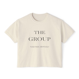 May include: A cream-coloured t-shirt featuring the words "THE GROUP" in black, centrally placed. Beneath this, in a smaller font, is "TOGETHER, OBVIOUSLY". The shirt has short sleeves and a crew neck.