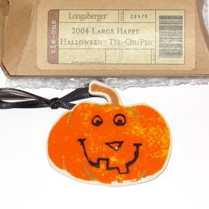 May include: A ceramic pumpkin-shaped tie-on pin with a smiling face, black ribbon, and a brown cardboard box. The pumpkin is orange with black facial features. The box has text that reads "2004 Large Happy Halloween Tie-On/Pin".