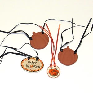 May include: Four Halloween-themed ceramic ornaments with black ribbon hangers. Two are pumpkin-shaped, one has the text "Happy Halloween" on a burlap background, and one has a painted image of pumpkins.