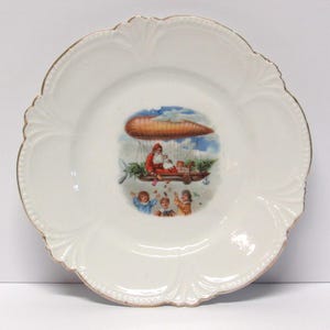 May include: A white ceramic plate with a scalloped edge and gold trim. The center features a colorful illustration of Santa Claus in an airship with children below. A decorative piece.