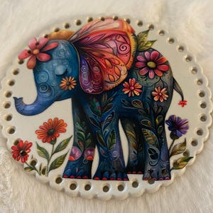 May include: A circular off-white base with a scalloped edge featuring evenly spaced holes around the perimeter. The center displays a blue elephant illustration adorned with colorful floral patterns, intricate swirls, and a large butterfly-like ear.