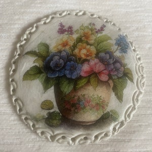May include: A white ceramic round plate with a decorative border. The plate features a painted image of a bouquet of flowers in a brown pot. The flowers are pink, yellow, blue, and purple.
