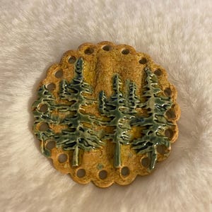 May include: A ceramic, circular decorative piece featuring a forest scene. The piece has a scalloped edge with evenly spaced holes. The trees are green and the background is a mix of yellow and brown.
