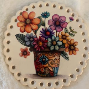 May include: A round, off-white ceramic disc with a scalloped edge featuring a series of evenly spaced holes around the perimeter. The center displays a colorful illustration of a bouquet of various flowers in a patterned, multi-colored pot.