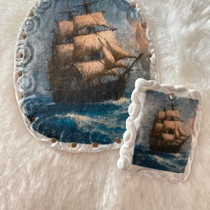 May include: Two decorative pieces featuring a ship at sea. One is oval-shaped with a detailed border, the other is rectangular. Both have a blue and white color scheme, depicting a ship sailing on rough waters.