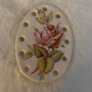 May include: Oval-shaped ceramic needle threader with a painted pink rose and green leaves. The threader has multiple small holes around the edge. The background is a soft, white, fuzzy texture.