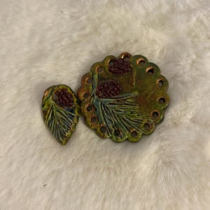 May include: Two ceramic pieces: a leaf-shaped pendant and a round, scalloped-edge brooch. Both feature a green and bronze glaze with a pinecone design in shades of brown. The brooch has small holes around the edge.