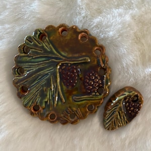 May include: Two ceramic pieces with a pine cone design. The larger piece is round with a scalloped edge and several holes. The smaller piece is leaf-shaped. The pieces are glazed in shades of green, brown, and gold.