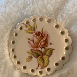 May include: A round, off-white ceramic coaster with a scalloped edge and evenly spaced holes. The center features a detailed illustration of a pink and orange rose with green leaves. A decorative item.