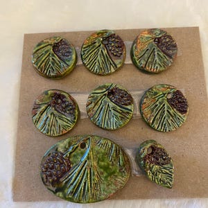 May include: A set of eight ceramic buttons in various shapes, including circles and an oval, featuring a green glaze with embossed pine cone and needle patterns. Each button has a single hole for attachment and a glossy, textured finish.