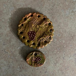 May include: Two round ceramic buttons with a pine tree design. The larger button has a scalloped edge and a brown and green glaze. The smaller button has a smooth edge and a brown and green glaze. Both buttons have red pine cones.