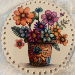 May include: A circular off-white ceramic plaque with a scalloped edge featuring evenly spaced holes around the perimeter. The center displays a colorful illustration of a bouquet of various flowers in a decorative pot with orange and blue swirls.