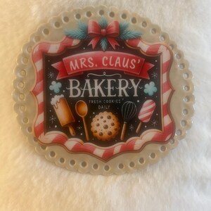 May include: A round decorative plate with a scalloped edge featuring a red and white candy cane border. The center displays a black sign reading MRS. CLAUS' BAKERY FRESH COOKIES DAILY, decorated with a red bow, rolling pin, spoon, cookie, and whisk.