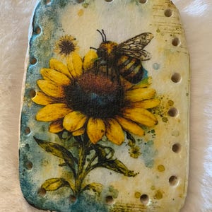 May include: Rectangular wooden plaque featuring a yellow sunflower with a bee. The sunflower has a dark brown center and green leaves. The background is a mix of yellow and blue. The plaque has small holes around the edges.