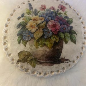 May include: Round, white decorative coaster with a floral bouquet design. The bouquet features yellow, blue, pink, and purple flowers in a brown vase. The coaster has a scalloped edge with small gold accents.