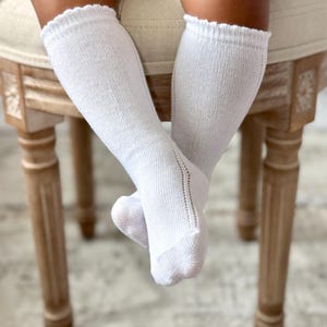 May include: White knee-high socks with a scalloped edge and a delicate openwork design on the side. The socks are made of a ribbed knit fabric and are shown on a light-colored background.