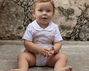 May include: A baby wearing a white collared shirt and a beige romper with lace detailing, sitting on a concrete surface. The background features a textured stone wall. The baby has blonde hair and is looking at the camera.