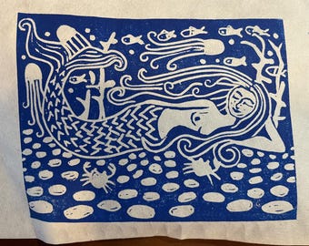 May include: A blue and white linocut print depicting a mermaid in a sea scene. The mermaid is surrounded by fish, jellyfish, and a crab. The print has a whimsical, artistic style, with a focus on flowing lines and organic shapes.