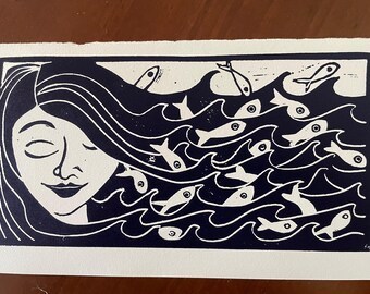 May include: A black and white linocut print depicting a woman's face with flowing hair, integrated with stylized waves and swimming fish. The artwork features a serene expression and a nautical theme.