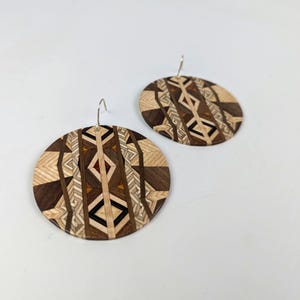 May include: A pair of round wooden earrings with a geometric pattern in shades of brown, beige, and black. The earrings are made of wood and have silver hooks.