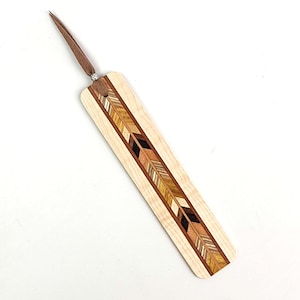 May include: A wooden bookmark with a decorative arrow design. The bookmark features a light wood base with a central inlay of various wood tones, including brown, black, and gold. A brown leather cord is attached at the top with a silver ring.