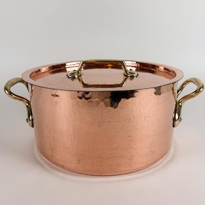 Puede incluir: Una olla redonda de cobre martillado con tapa a juego y dos asas laterales de latón curvado. La olla presenta un acabado metálico pulido con una superficie texturizada, y la tapa incluye un asa central de latón para facilitar su uso.