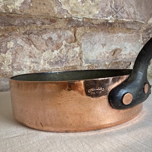 May include: A round copper sauté pan with a dark metal handle attached by two copper rivets. The side of the pan features a stamped mark reading GAILLARD PARIS. The interior is lined with a silver-colored metal, and the exterior shows a polished copper finish with minor surface scratches.