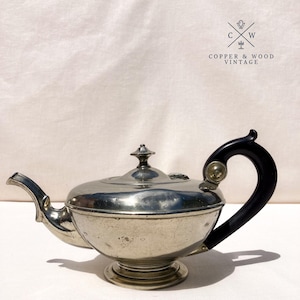 May include: Vintage silver teapot with a black handle and a decorative finial on the lid. The teapot has a spout and sits on a round base. The handle is curved and attached to the body with a small gold-colored accent. The text "COPPER & WOOD VINTAGE" is in the upper right corner.