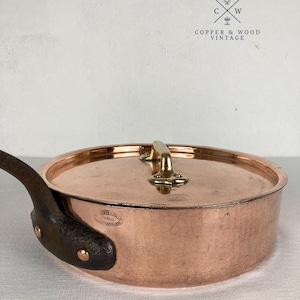 Puede incluir: Una sartén redonda de cobre con tapa, que presenta un mango de metal oscuro sujeto con remaches de cobre. La tapa tiene un asa de latón y una marca estampada que dice "CUVILLERIE PARIS". El logotipo "COPPER & WOOD VINTAGE" es visible.