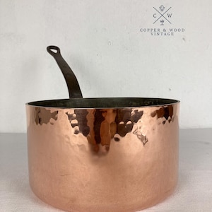 Könnte beinhalten: Ein gehämmertes Kupfer-Kochgeschirr mit einem dunklen Metallgriff, der seitlich absteht. Die Außenfläche weist eine reflektierende, strukturierte Oberfläche auf. Das Logo COPPER & WOOD VINTAGE mit einem gekreuzten Utensilien-Symbol ist sichtbar.